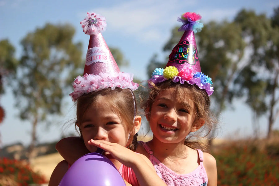 Two girls smiling in pink party hats and holding a purple balloon